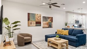 Een moderne woonkamer met een blauwe bank, een bruine fauteuil, een houten salontafel en een plafondventilator. Er is een keuken met witte kasten en een magnetron.
