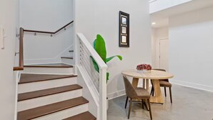 Een trap met houten treden en witte leuningen, een ronde houten tafel met stoelen, en een groene plant aan de muur.