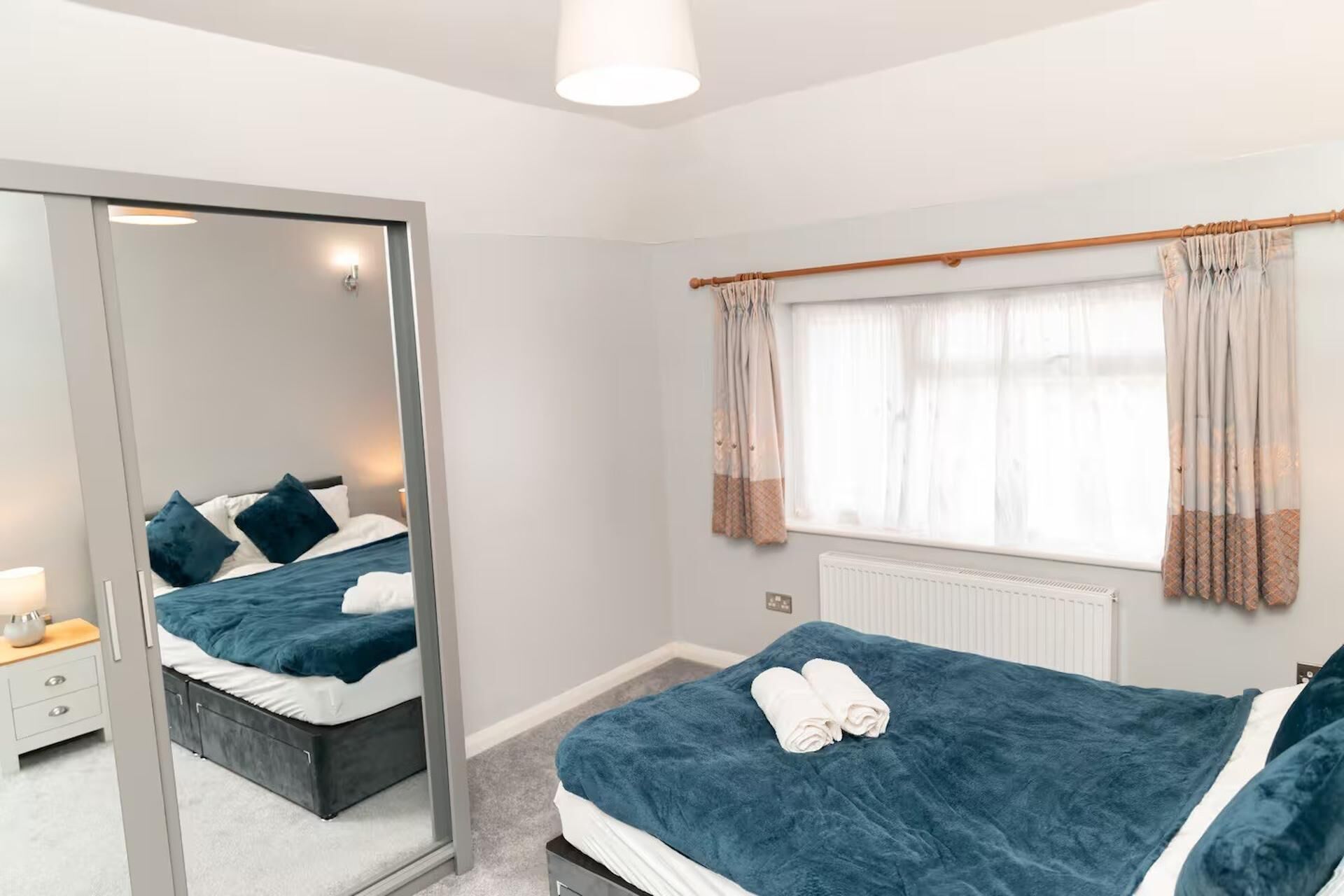 A bedroom with a large mirror, two beds with blue bedding, a bedside table with a lamp, and a window with curtains.