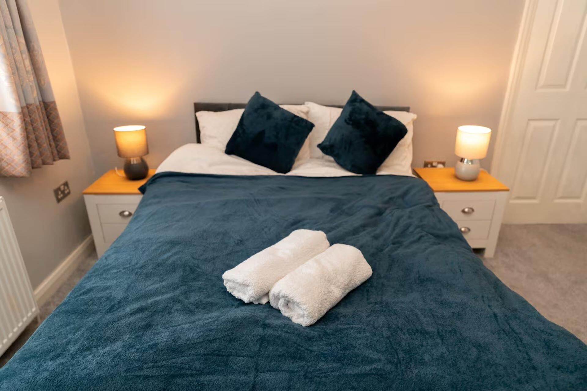 A neatly made bed with a dark blue bedspread, two white towels, and two dark blue pillows. There are two bedside lamps on wooden nightstands.