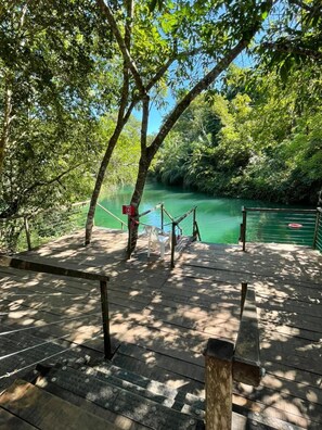 A wooden deck with a rope barrier, a bench, and a view of a calm river surrounded by dense greenery. - Camping Rio Formoso LTDA (Bonito)