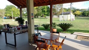 An outdoor patio with a table and chairs, surrounded by potted plants and a view of a grassy area with trees and a building in the background.