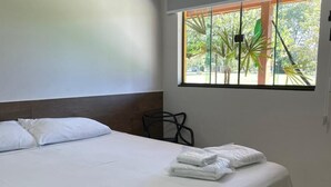 A hotel room with a bed, a window with a view of greenery, a chair, and a wall-mounted air conditioning unit.