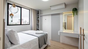 A modern bedroom with a bed, a window with a city view, a built-in shelf, and a mirror with a plant. - Fully Equipped Apartments in a Building With a Pool, 400m From Picarras Beach - Bacs (Picarras)