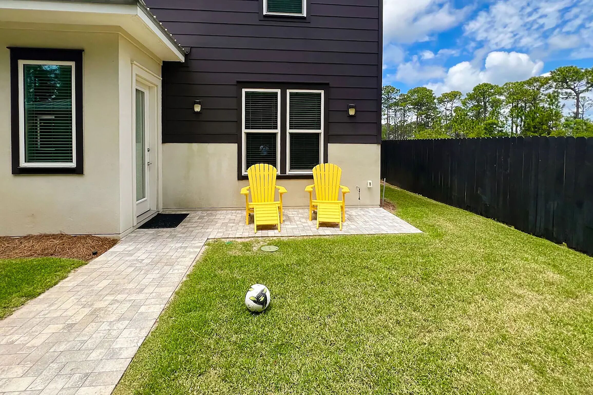 Une maison avec une terrasse dallée, deux chaises jaunes et un ballon de foot sur la pelouse.
