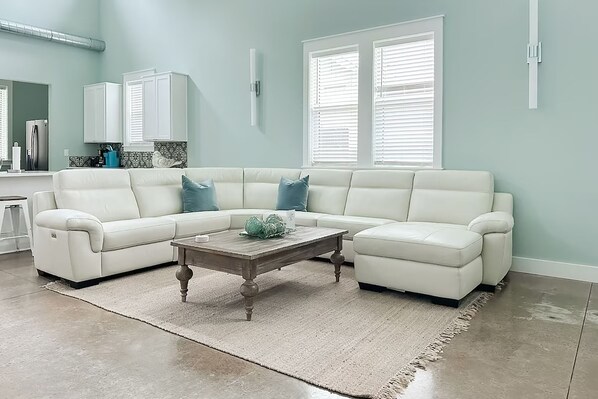 A modern living room with a sectional sofa, a wooden coffee table, and a kitchen area in the background. - Down da Road (Miramar Beach)