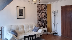 Ein Wohnzimmer mit einem weißen Sofa, einem Esstisch mit Tellern und Gläsern, einer Kronleuchter, einem Kleiderständer und einer gemusterten Tapete.