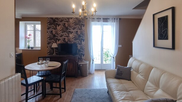 Ein Wohnzimmer mit einem weißen Sofa, einem Esstisch mit Stühlen, einer Chandelüster und Blick ins Freie durch ein Fenster.