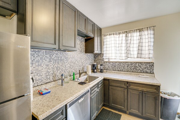 Una cocina con refrigerador de acero inoxidable, salpicadero de azulejos, un lavamanos y una ventana con cortinas a cuadros.
