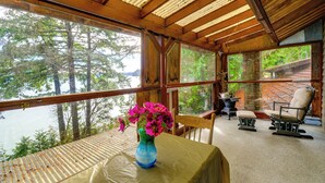 Une cabane en bois dotée d’une grande fenêtre donnant sur un lac et une forêt. À l’intérieur, on trouve une table ornée d’un vase de fleurs et deux chaises.