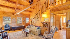 A spacious log cabin with a large living area featuring a sofa, chairs, and a coffee table. A staircase leads to an upper level, and the cabin has large windows. - On-Site Trails, Pond! Rustic Harpers Ferry Cabin (Harpers Ferry)