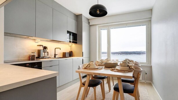 Eine moderne Küche mit grauen Schränken, einem Holzespeisetisch gedeckt mit Tellern und Blick auf einen See durch ein großes Fenster.