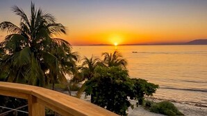 Une terrasse en bois donnant sur une plage bordée de palmiers, avec un coucher de soleil en toile de fond.