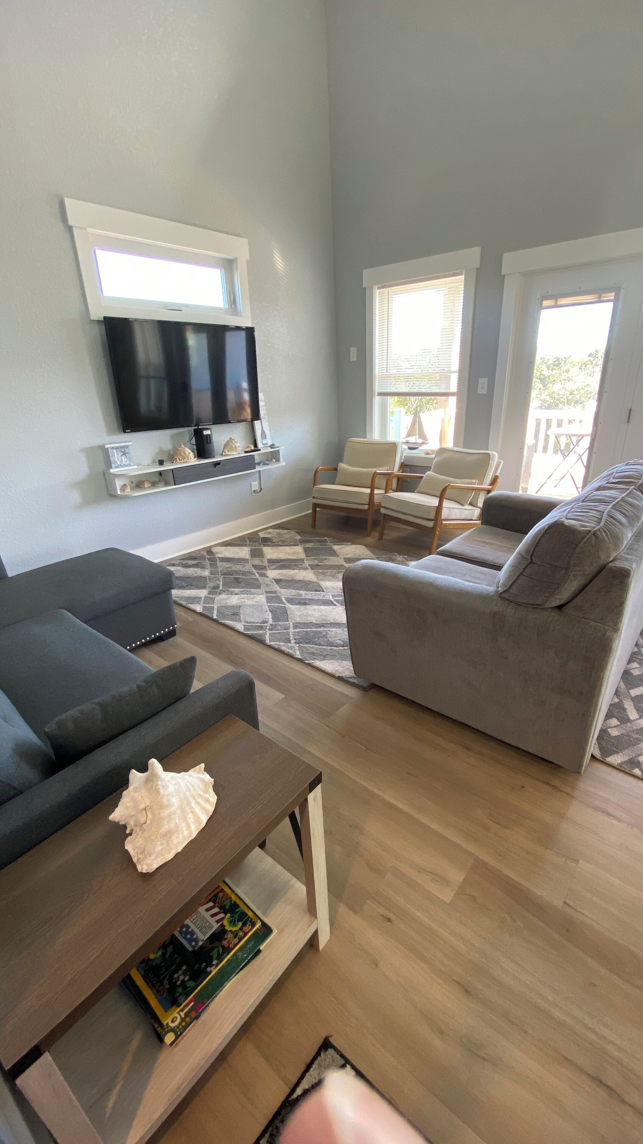 A living room with a flat-screen TV mounted on the wall, a grey sofa, a coffee table with a book, and a patterned rug.