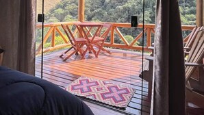 A room with a bed, a view of a wooden deck, and a balcony with chairs and a table. - Chalé Recanto da Mata - Romantic chalet with mountain views and a hot tub. (Sapucaí-Mirim)