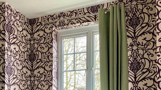 Una habitación con papel tapiz estampado, una ventana con cortinas verdes, un banco blanco y un pequeño otoman blanco con un florero de flores.