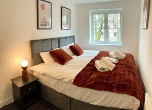 A neatly made bed with white linens, brown pillows, and a brown throw blanket. There is a nightstand with a lamp, and a window with a view of trees. - Comfy & Charming Flat Family & Corporate Friendly (Liverpool)