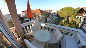Un balcón con una mesa redonda, dos sillas, un paraguas rojo y vistas a una zona residencial con casas y árboles.