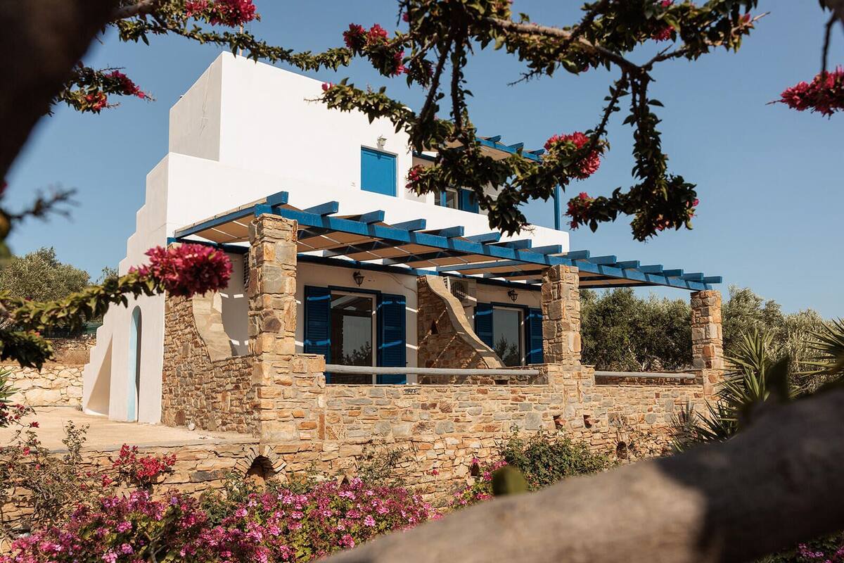 A white building with blue shutters and a stone foundation, surrounded by pink flowers and greenery.