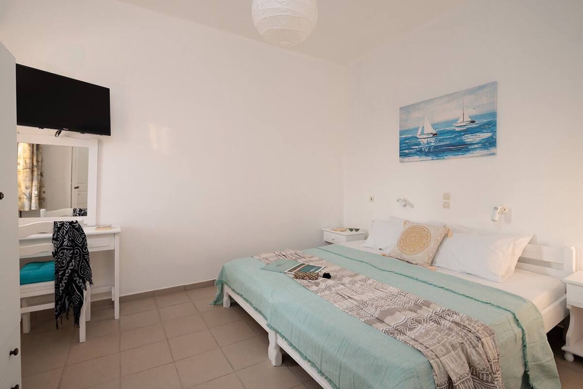 A bedroom with a bed, a bedside table, a nightstand, a TV mounted on the wall, and a painting of a sailboat.