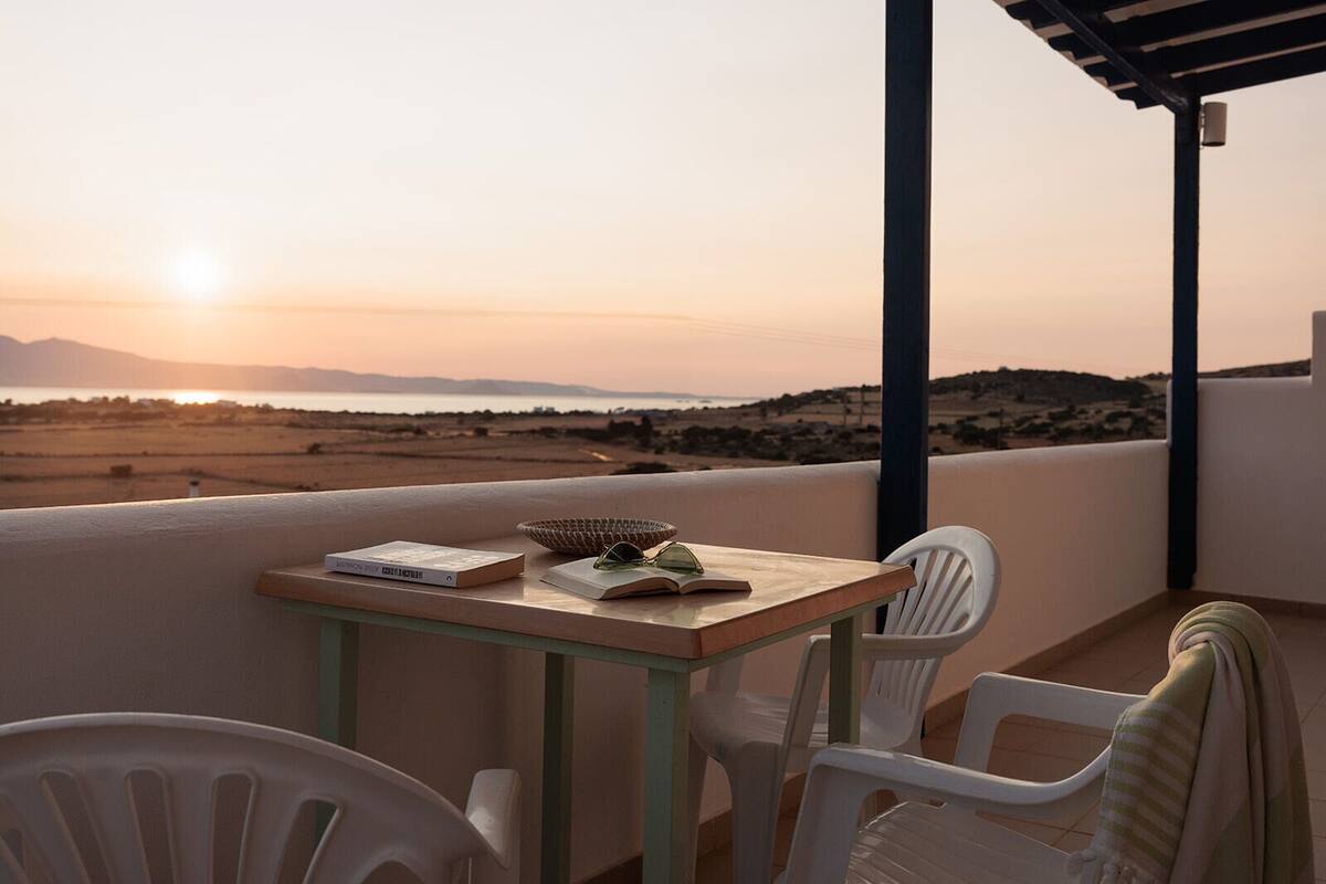 A balcony with a table, chairs, and a view of a sunset over a landscape.