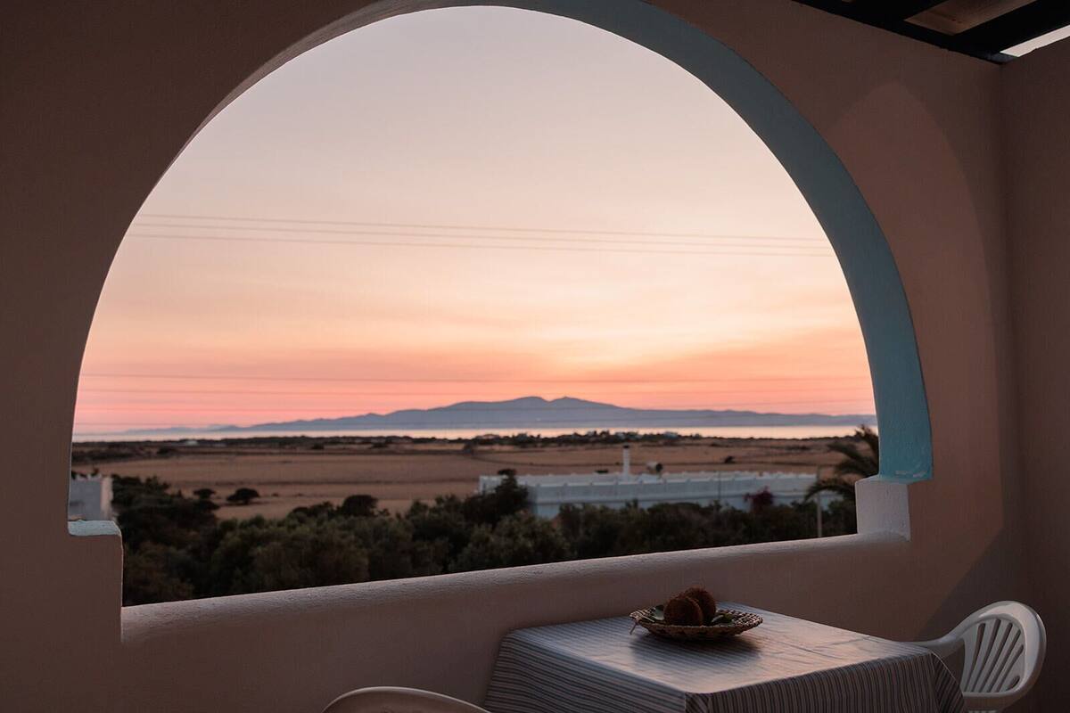 A balcony with a view of a sunset over a landscape, a table with a plate, and a chair.