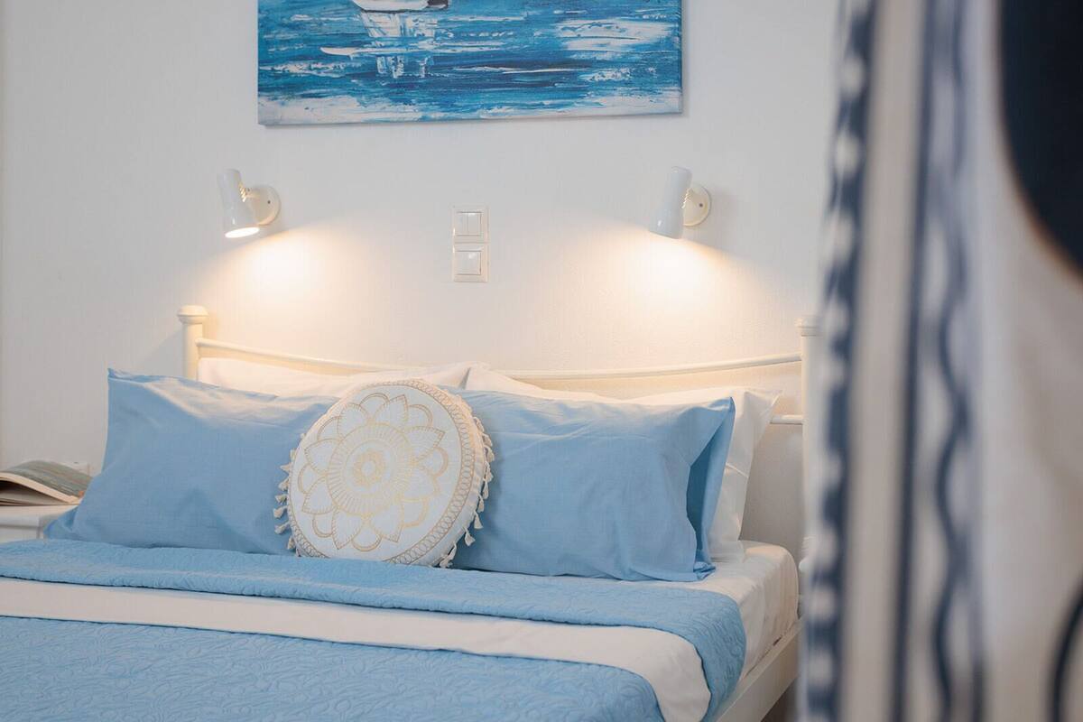 A neatly made bed with blue bedding and a decorative pillow, a painting of a blue seascape above it, and wall-mounted lights on either side.