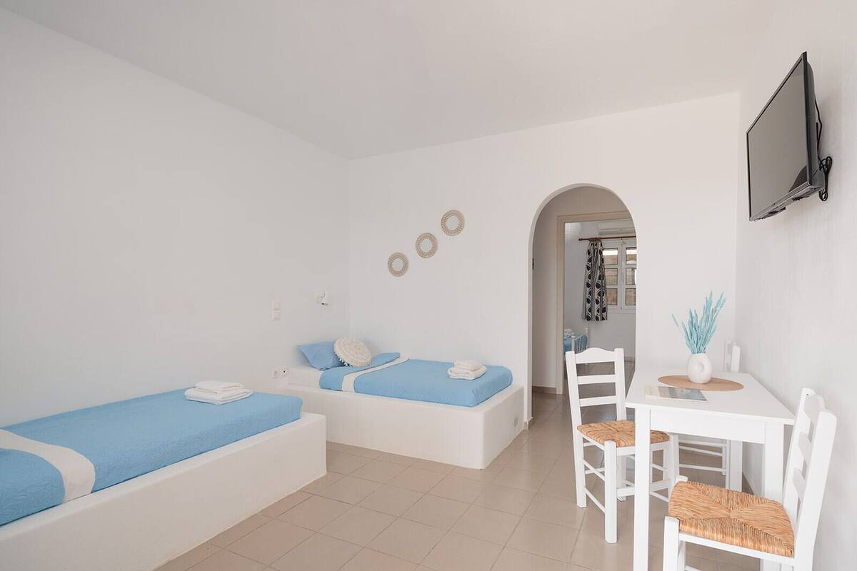 A modern, minimalist room with two single beds, a white dining table with chairs, and a flat-screen TV mounted on the wall.