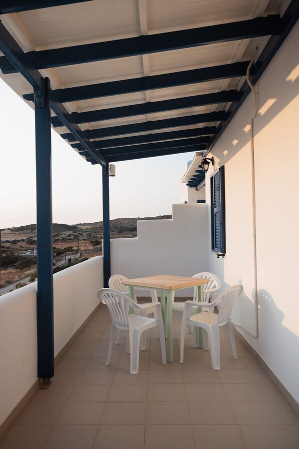 Une terrasse extérieure couverte, avec une table et des chaises, donnant sur la campagne, et un bâtiment aux volets bleus.