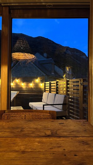 Een houten terras met een bank en tafel, sfeerverlichting en uitzicht op een berg in de schemering.