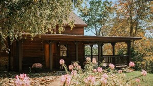 Uma cabana de madeira com varanda coberta, rodeada por um jardim com flores cor-de-rosa e um caminho de pedra.