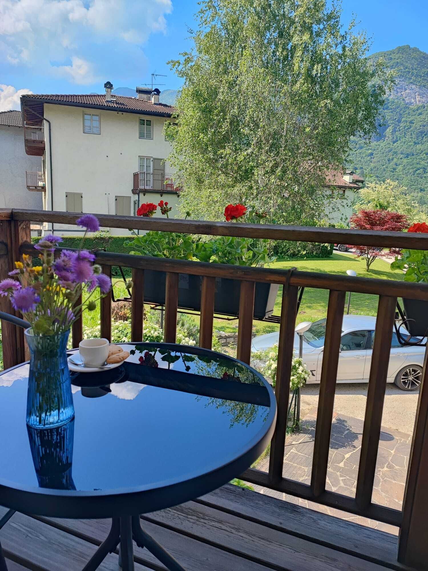 En balkon med et rundt bord, en vase med blomster og udsigt til et bjerg samt en bil i baggrunden.