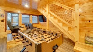 Una habitación con piso de madera, una mesa de fútbol de mesa, una escalera y dos sillas negras.