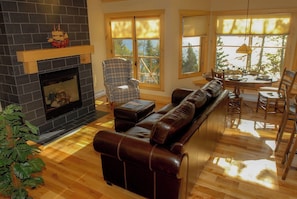 Una sala de estar con chimenea, un sofá de cuero, una mesa de comedor con sillas y ventanas grandes con persianas.