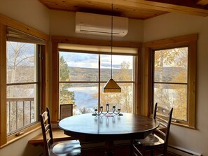A round dining table with chairs, a hanging pendant light, and a view of a lake through large windows. - Magnificent Luxury Condo at Lake Pohénégamook (Pohénégamook)