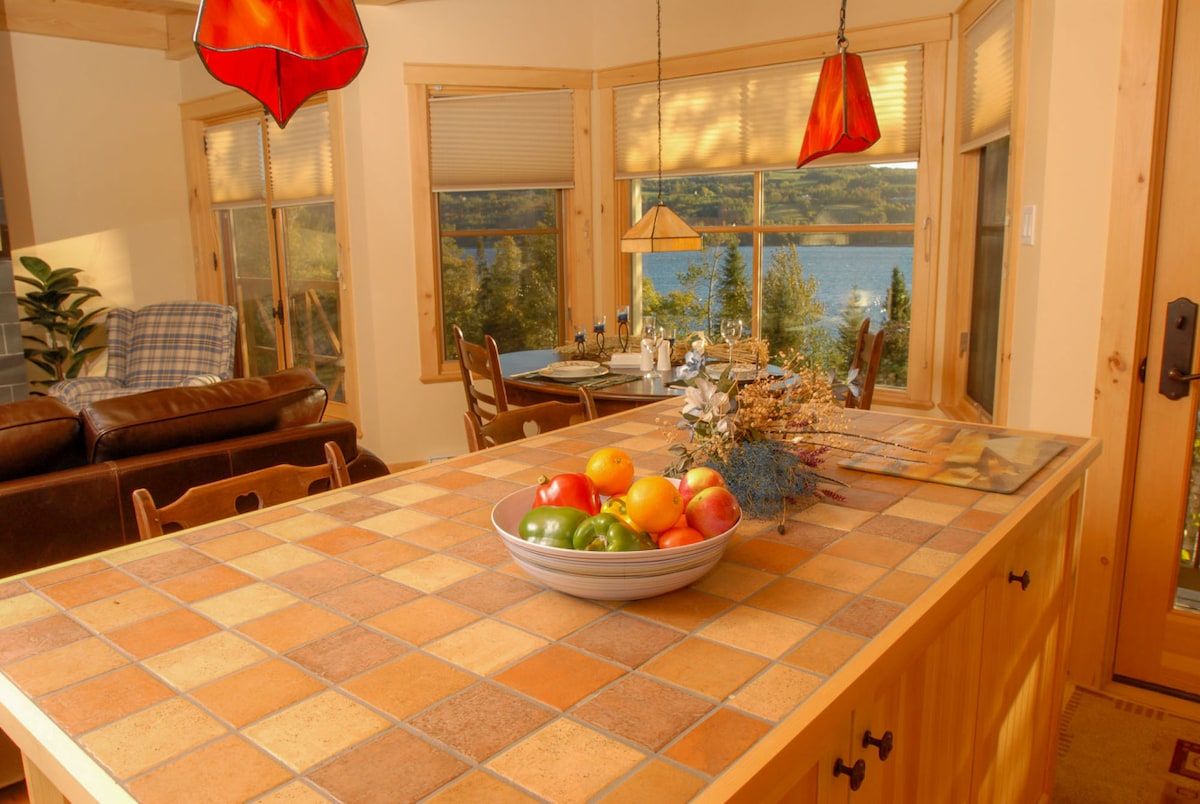 A kitchen with a checkered table, a bowl of fruit, and a view of a lake.