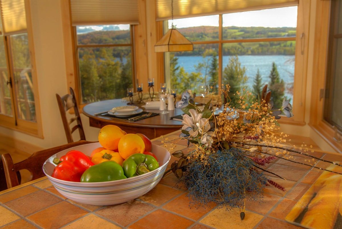 Una mesa de comedor con un tazón de fruta y un arreglo floral, con vistas a un lago pintoresco.