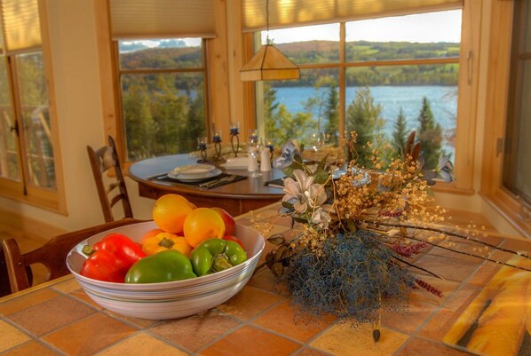 A dining table with a bowl of fruit and a floral arrangement, set against a backdrop of a scenic lake view.