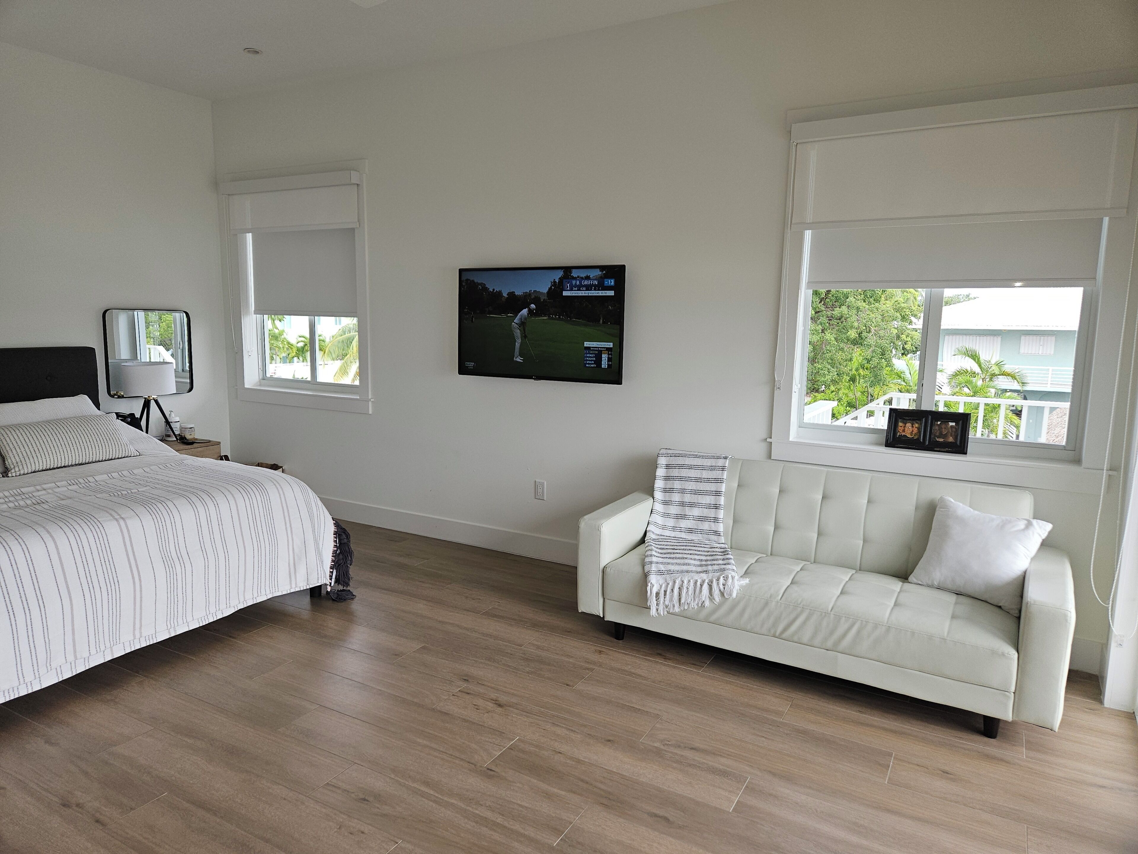 A bedroom with a bed, a nightstand, a TV on the wall, and a window with a view of greenery.