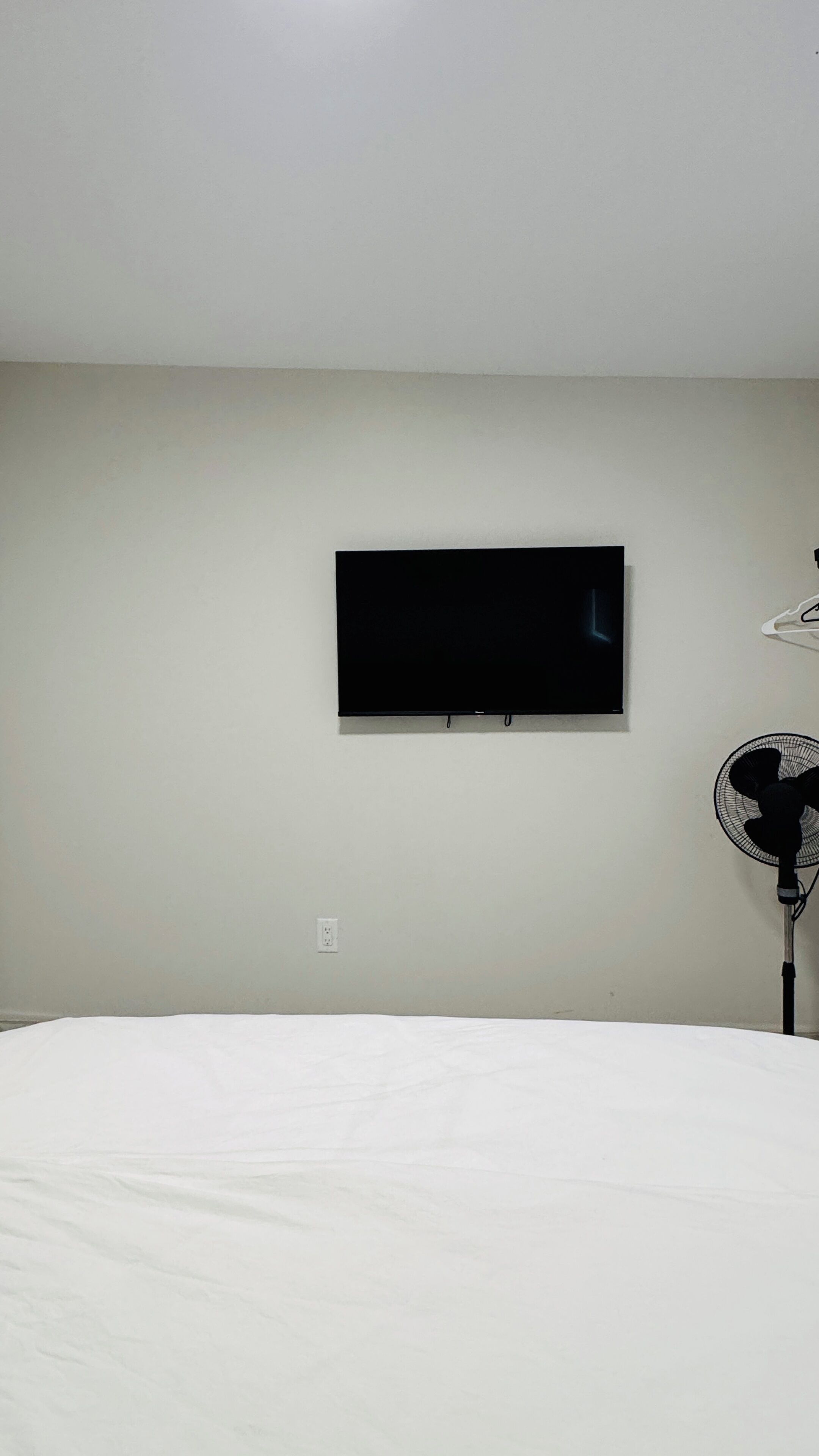 A bedroom with a bed, a wall-mounted TV, and a standing fan.