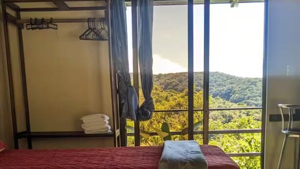 A room with a bed, a small table, and a chair. There is a window with a view of greenery and hills. - villa sunset (Santa Elena)