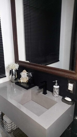 Un baño con un lavamanos blanco, grifo negro y un espejo encima. Hay un jarrón con flores al lado izquierdo del lavamanos.