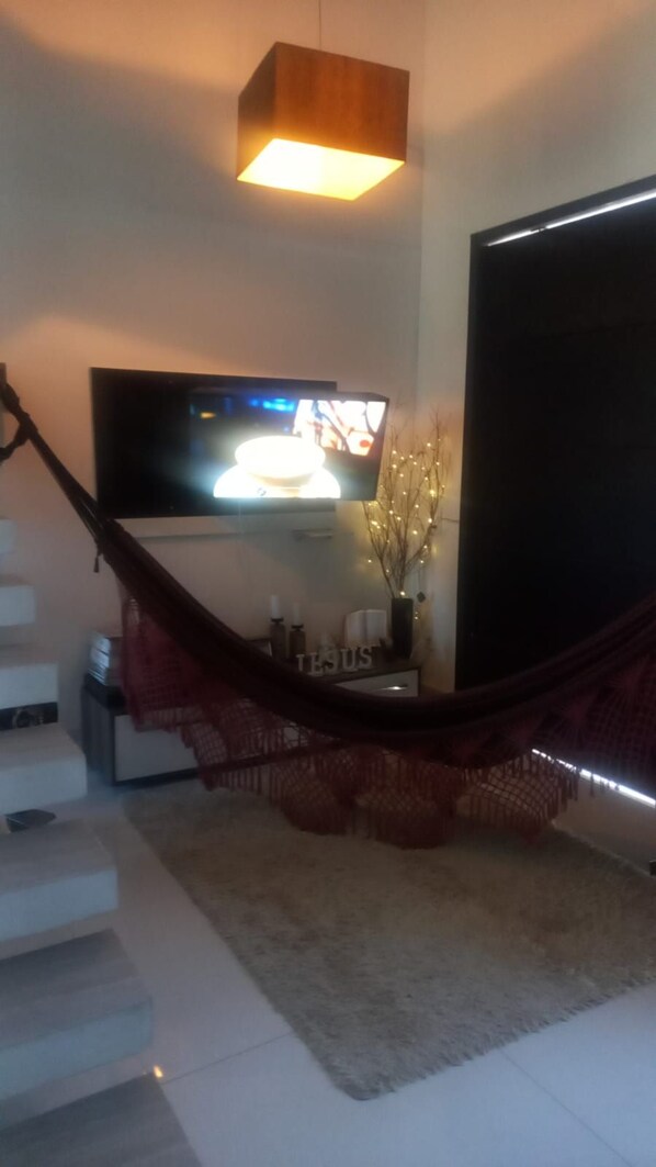 A living room with a TV mounted on the wall, a hammock, and a staircase. - Pousada Flor de Lis Homestay (Volta Redonda)