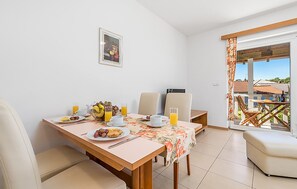 Ein Essbereich mit einem Tisch, der für das Frühstück gedeckt ist, einschließlich Früchten, Saft und einer Kaffeetasse. Es gibt zwei Stühle, ein Sofa und einen Ausblick nach draußen durch ein Fenster mit Vorhängen.