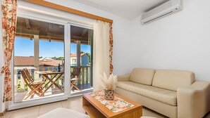 A living room with a beige sofa, a wooden coffee table with a floral table runner, and a view of a balcony with a table and chairs. - Nice apartment in Palit with swimming pool (Palit)
