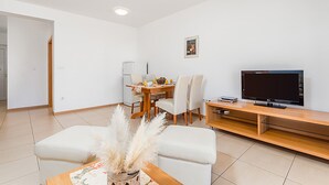 A modern living room with a dining area, a flat-screen TV, and a small table with decorative items. - Nice apartment in Palit with swimming pool (Palit)