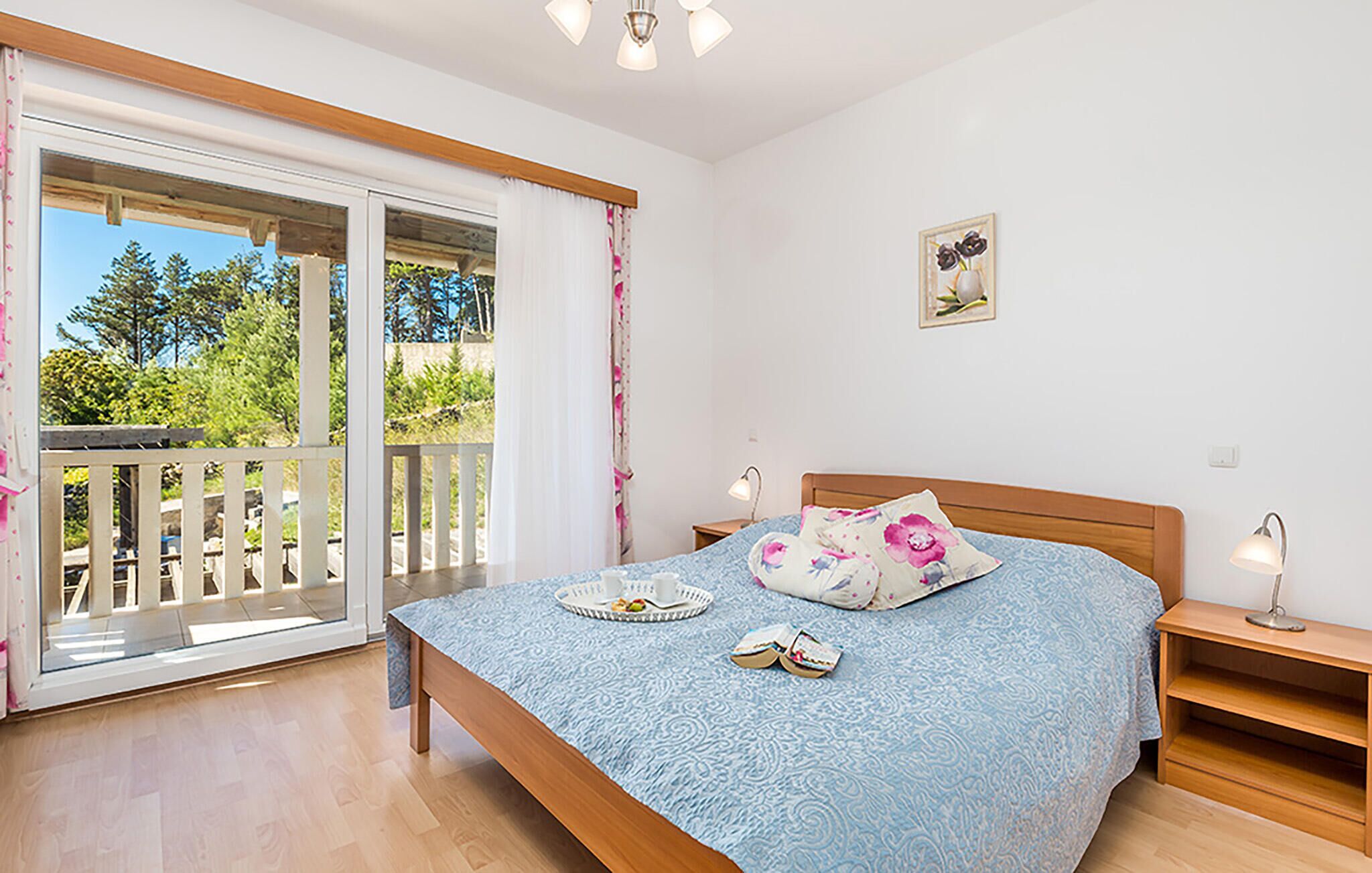 A bedroom with a bed, two bedside tables, a nightstand, a window with curtains, and a view of greenery outside.