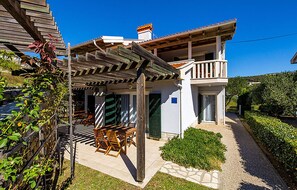 Una casa de dos pisos con balcón, un porche cubierto con mobiliario exterior y un jardín cuidado.