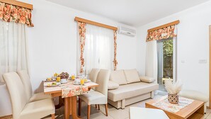 Een moderne woonkamer met een eettafel, bank en salontafel. Er is een raam met gordijnen, een airconditioner en uitzicht op groen.