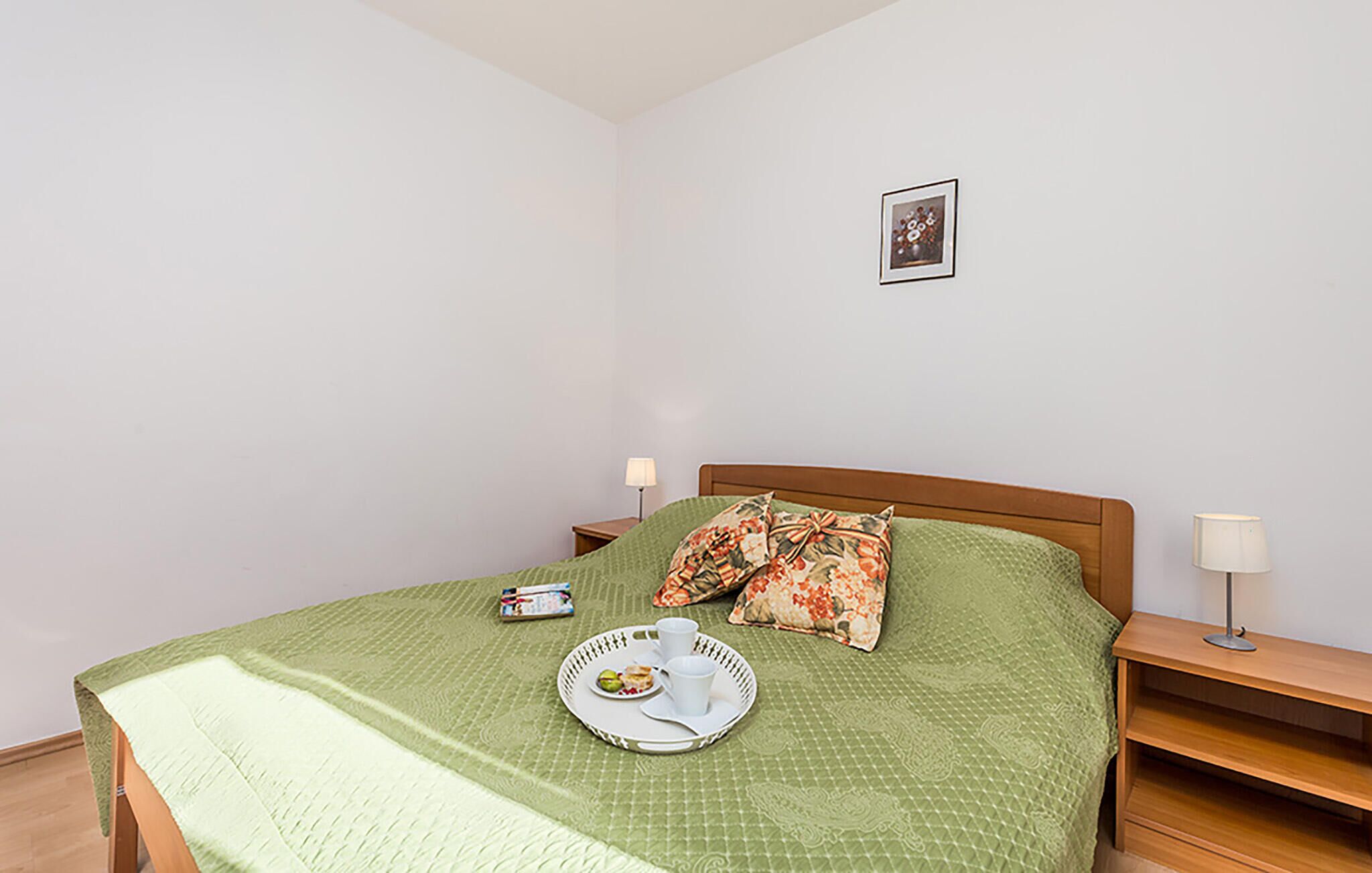 A neatly made bed with a green quilt, a floral pillow, and a tray with a plate of food and a cup. There are two bedside tables with lamps, and a framed picture on the wall.